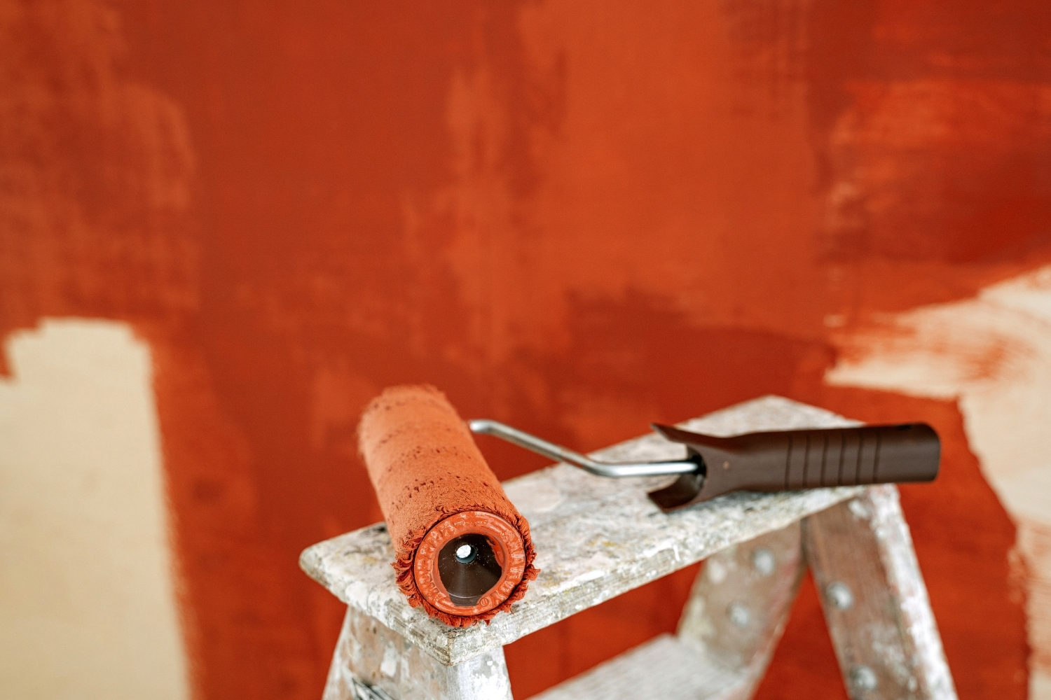 Paint roller resting on a step ladder with an unfinished wall behind, being painted in a red-orange colour.